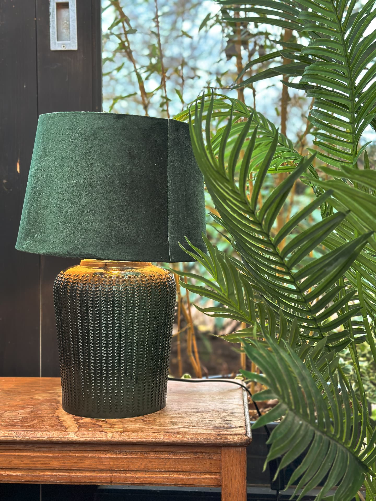 Green velvet lamp on a wooden table beside palm foliage at Midpines